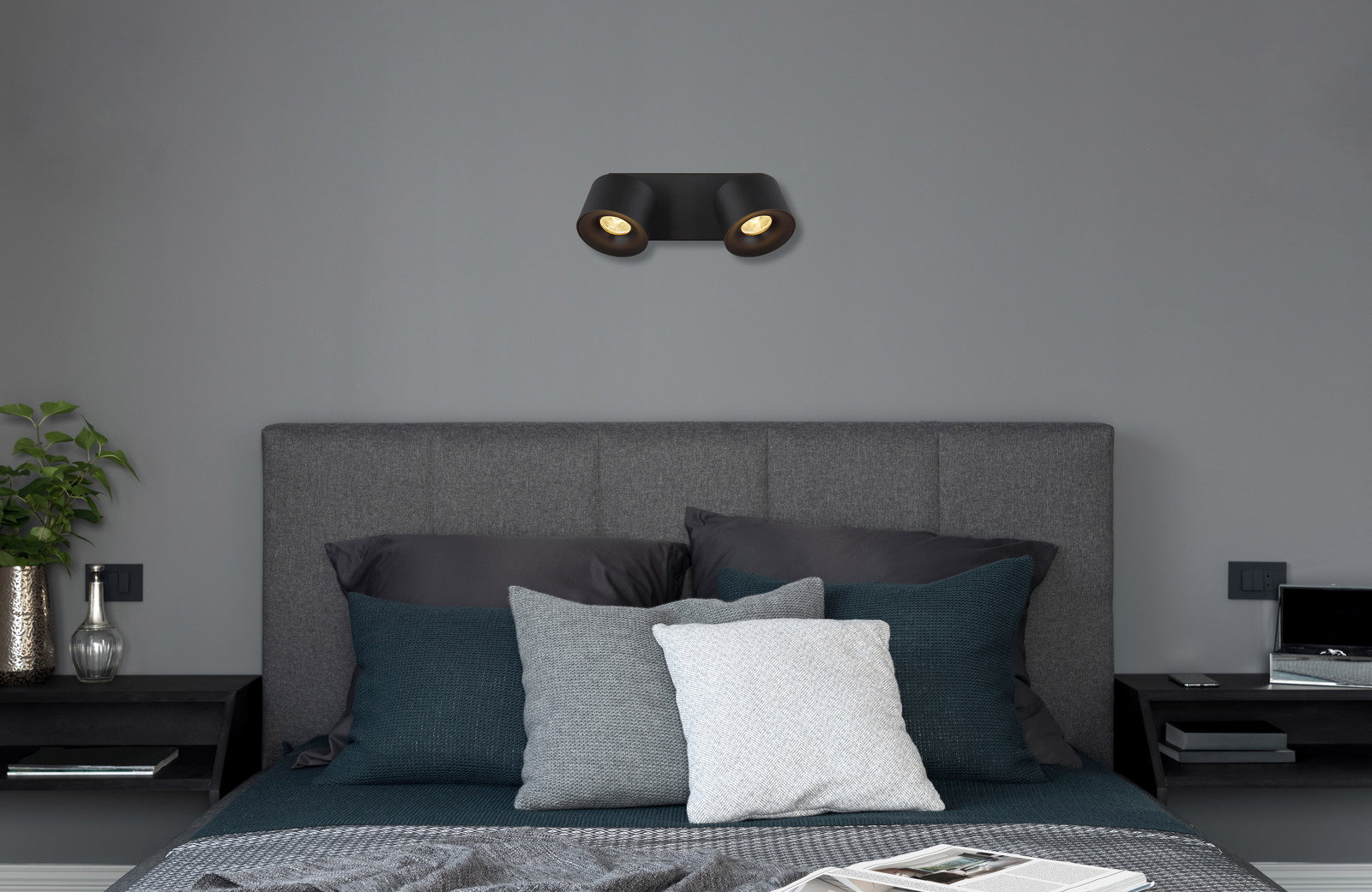 Schwarzer LED Strahler mit zwei Leuchten, an der Wand über einem grauen Bett montiert, seitliche Perspektive