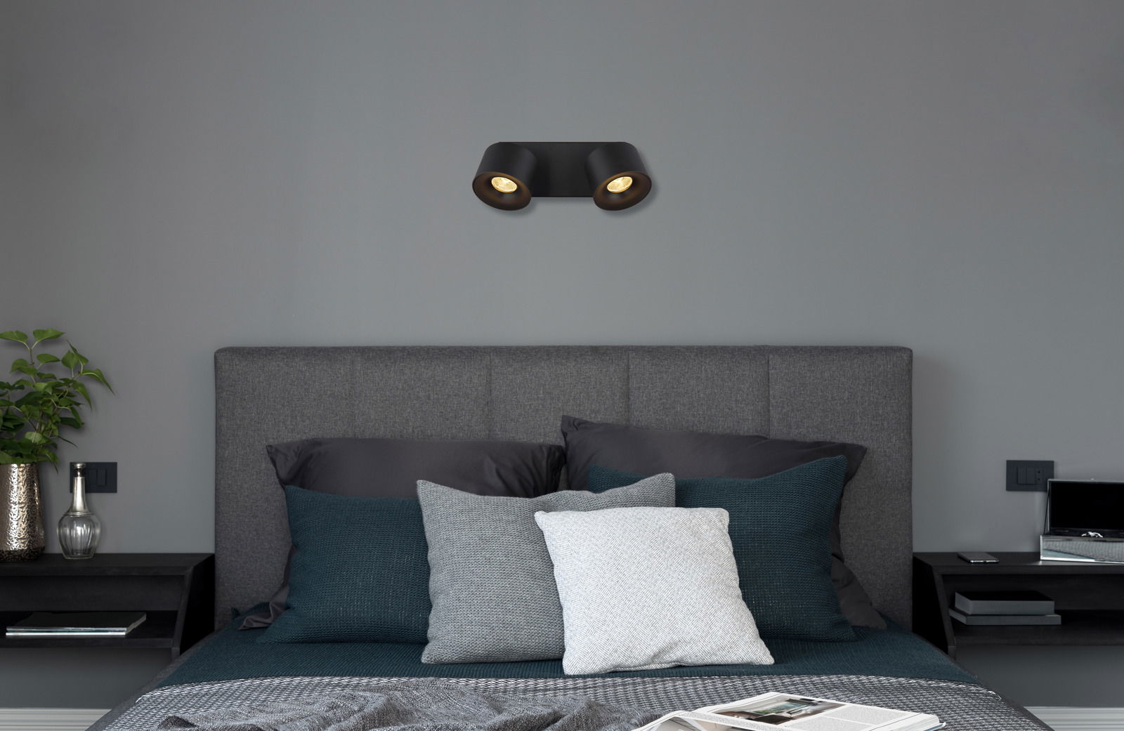 Schwarzer LED Strahler mit zwei Leuchten, an der Wand über einem grauen Bett montiert, seitliche Perspektive