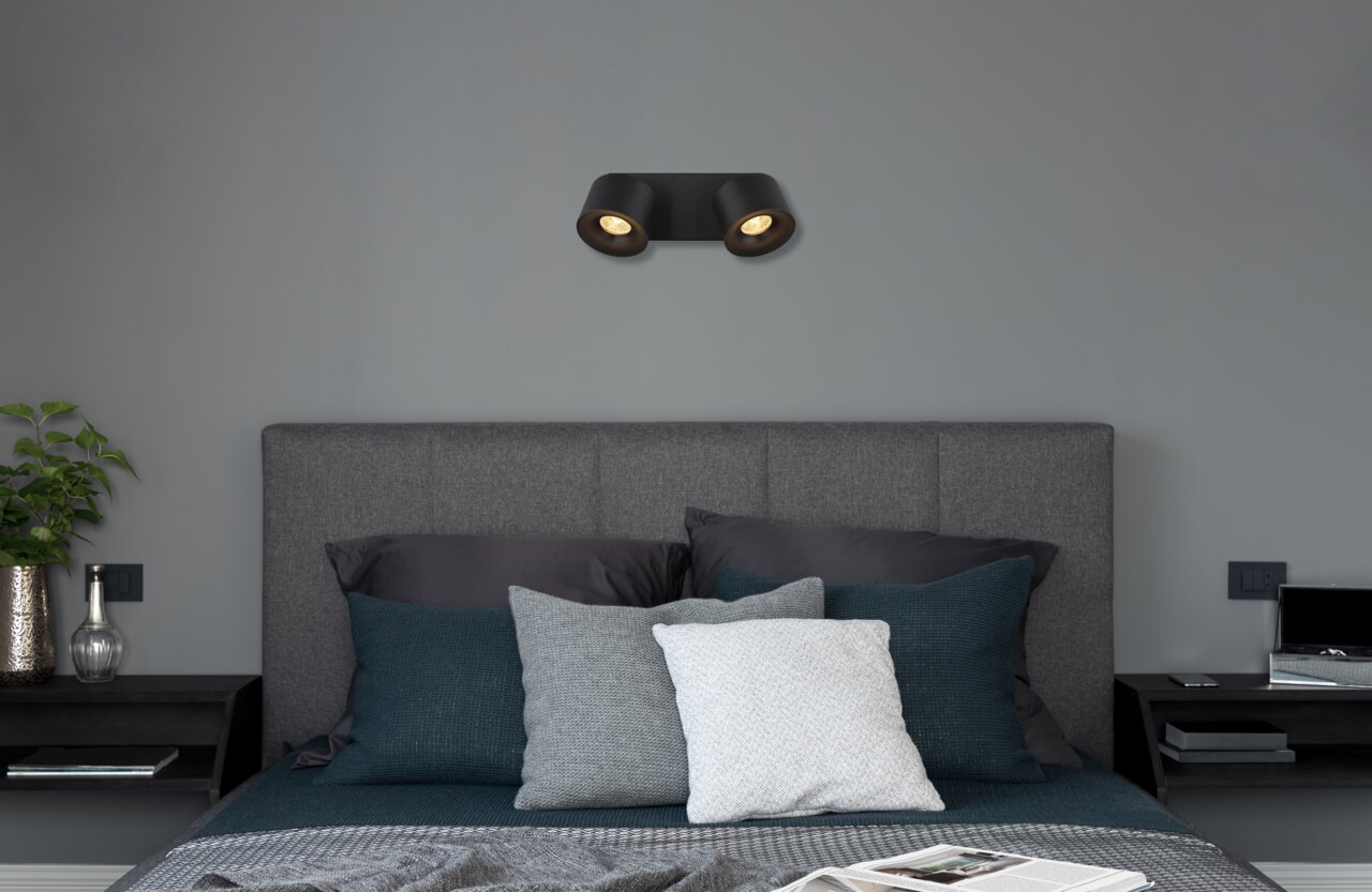 Schwarzer LED Strahler mit zwei Leuchten, an der Wand über einem grauen Bett montiert, seitliche Perspektive