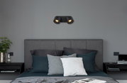 Schwarzer LED Strahler mit zwei Leuchten, an der Wand über einem grauen Bett montiert, seitliche Perspektive