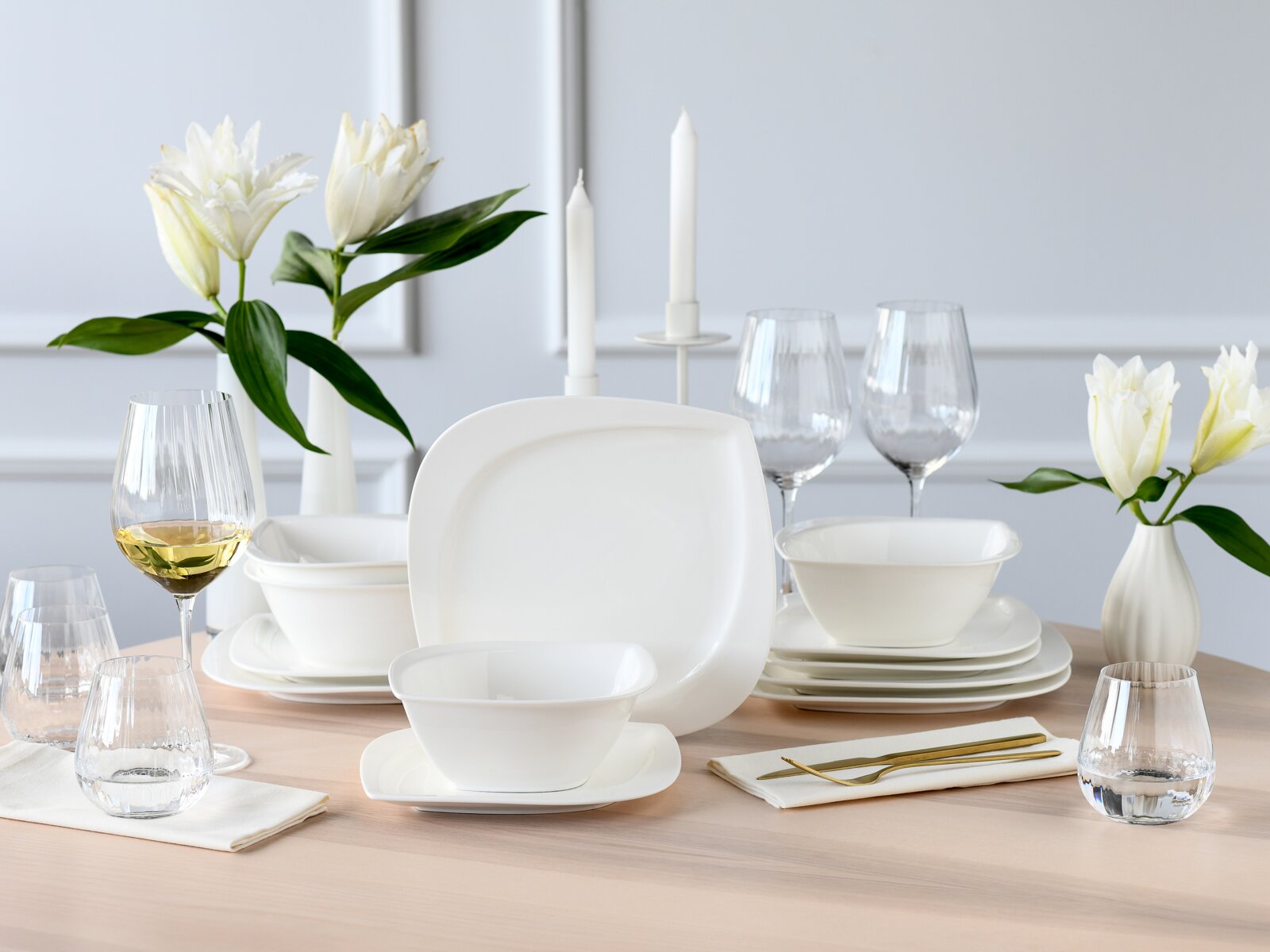 Creatable Tellerset 12-tlg MELBOURNE Modernes weißes Porzellan-Tafelservice mit eckig abgerundeten Tellern und Schalen, dekoriert mit Gläsern, Kerzen und weißen Blumen auf Holztisch, frontal aus leicht erhöhter Perspektive