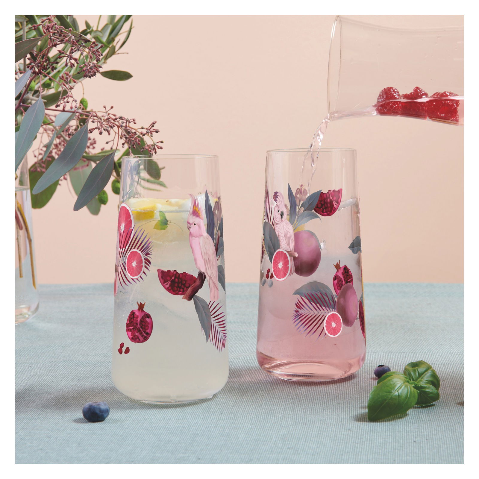 Zwei dekorative Gläser mit exotischen Vogel- und Fruchtmotiven, gefüllt mit Getränken, auf einem Tisch mit Pflanzen im Hintergrund. Seitliche Perspektive.