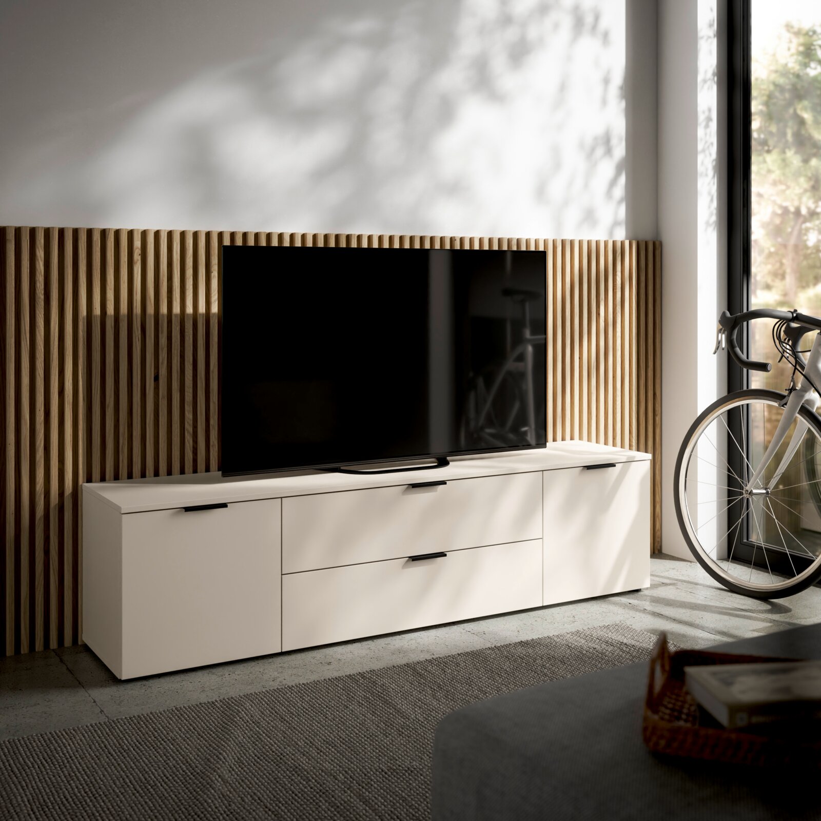 Modern tv-lowboard in een lichte kleur met lades en deuren, gezien vanaf de voorkant links, naast een grote tv en een fiets.