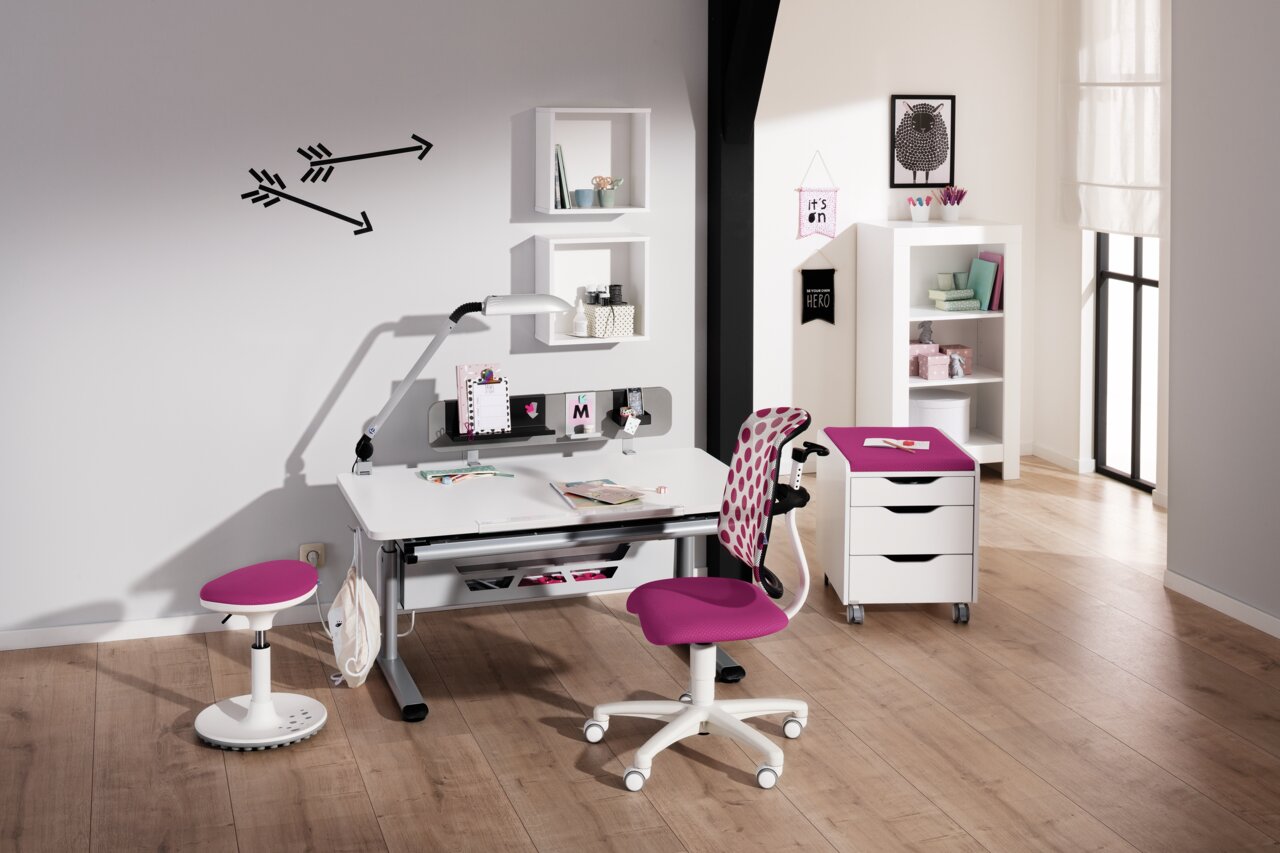 Moderner Schreibtisch mit weißer Tischplatte und grauen Beinen, umgeben von einem pink gepolsterten Drehstuhl und einem Hocker. Im Hintergrund sind Regale und Dekorationen zu sehen. Perspektive: Frontalansicht.