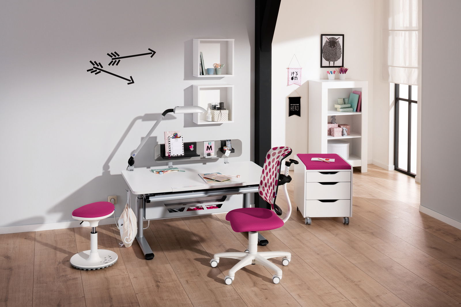 Moderner Schreibtisch mit weißer Tischplatte und grauen Beinen, umgeben von einem pink gepolsterten Drehstuhl und einem Hocker. Im Hintergrund sind Regale und Dekorationen zu sehen. Perspektive: Frontalansicht.