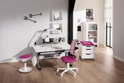 Moderner Schreibtisch mit weißer Tischplatte und grauen Beinen, umgeben von einem pink gepolsterten Drehstuhl und einem Hocker. Im Hintergrund sind Regale und Dekorationen zu sehen. Perspektive: Frontalansicht.