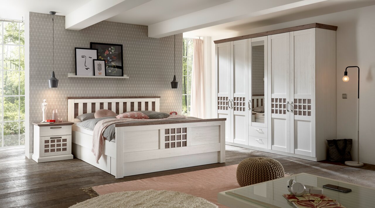 Schlafzimmeransicht mit einem modernen, weißen Kleiderschrank mit fünf Türen, einem Doppelbett mit dekorativen Kissen und einer Nachtkommode, aus der Perspektive von der Zimmerecke aus fotografiert.