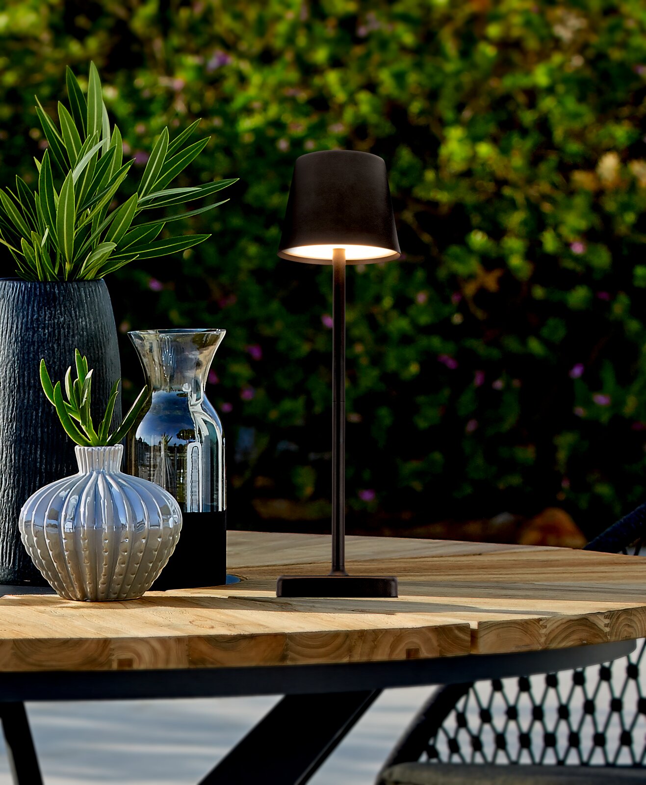 Ambia Garden Solarleuchte MOBILE Elegante Solarleuchte auf einem Holztisch im Freien, umgeben von dekorativen Vasen und Pflanzen, seitliche Perspektive.