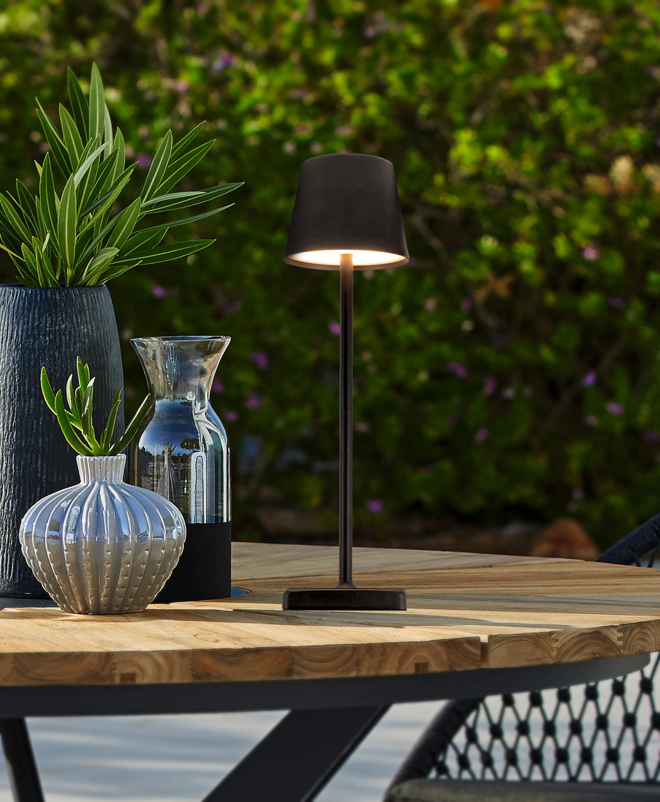 Elegante Solarleuchte auf einem Holztisch im Freien, umgeben von dekorativen Vasen und Pflanzen, seitliche Perspektive.