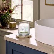Brandende Yankee Candle Clean Cotton geurkaars met twee lonten in een glazen pot van 368 g, vanuit schuin vooraanzicht naast een vrijstaand bad