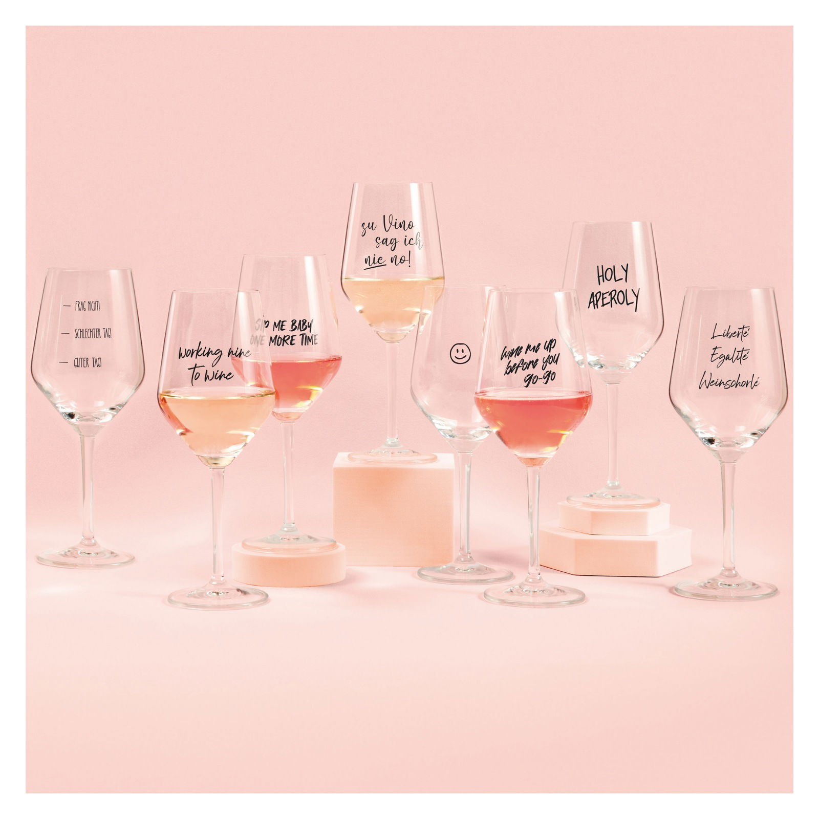 Eine Sammlung von Weingläsern mit humorvollen Sprüchen, gefüllt mit Roséwein, auf einem rosa Hintergrund. Frontale Perspektive.