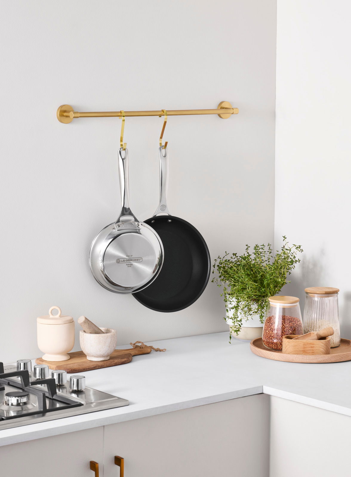 Zwei Pfannen aus Edelstahl, eine mit Antihaftbeschichtung, hängen an einer goldenen Stange in einer modernen Küche. Perspektive von der Seite.