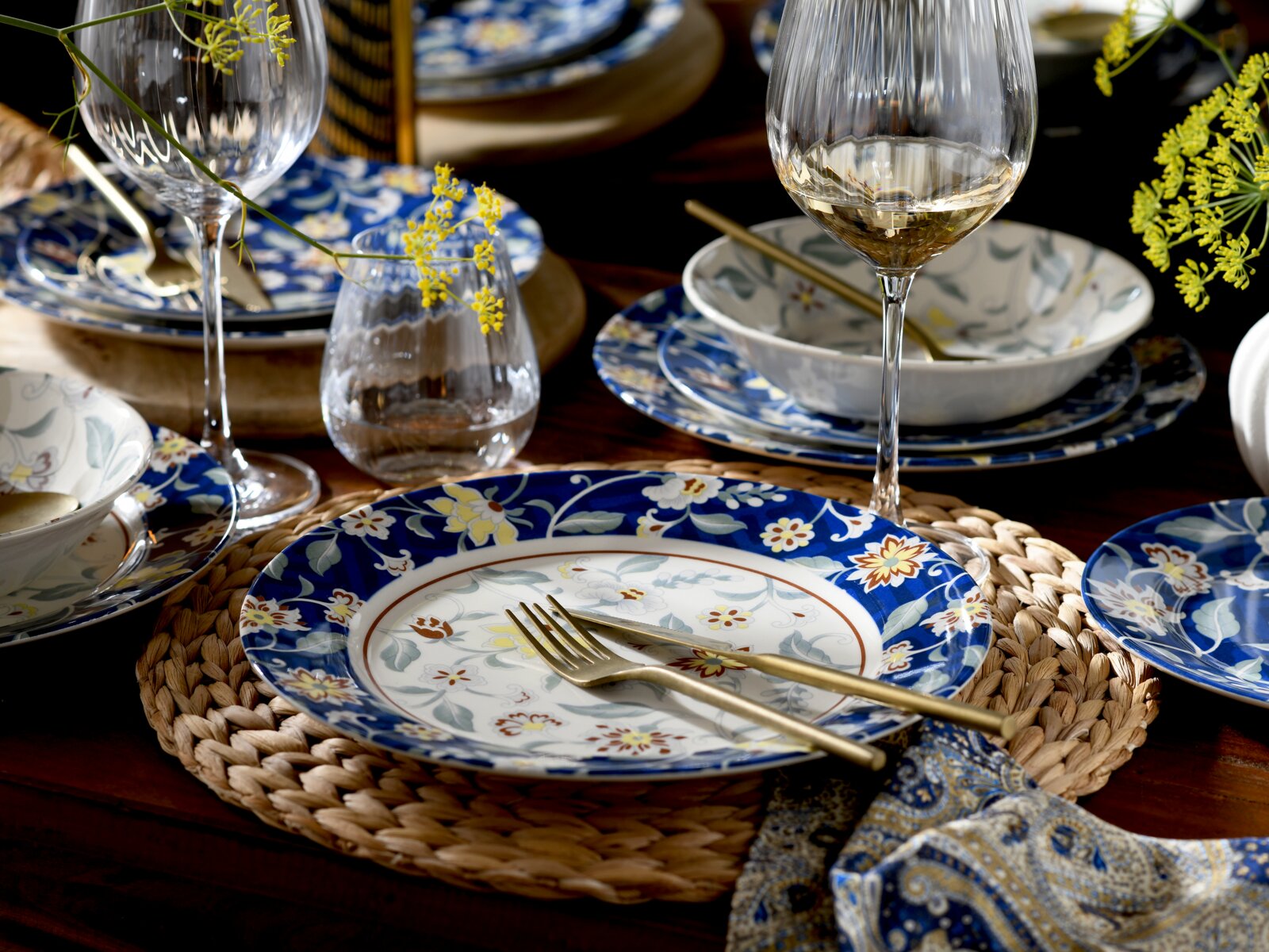 Schräg von oben fotografierte, stilvoll gedeckte Tafel mit blau-weißem, floral gemustertem Geschirr, goldfarbenem Besteck, Weingläsern und gelben Blüten auf dunklem Holztisch.