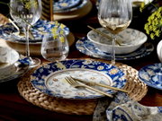 Schräg von oben fotografierte, stilvoll gedeckte Tafel mit blau-weißem, floral gemustertem Geschirr, goldfarbenem Besteck, Weingläsern und gelben Blüten auf dunklem Holztisch.