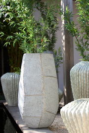 Großer, moderner Pflanztopf aus Beton mit geometrischem Design, gefüllt mit grünen Pflanzen, in einer Außenumgebung. Perspektive von vorne.