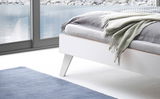 Nahaufnahme eines modernen weißen Bettgestells mit schrägen Beinen, im Vordergrund eine blaue Decke und im Hintergrund ein Fenster mit Blick auf einen See.