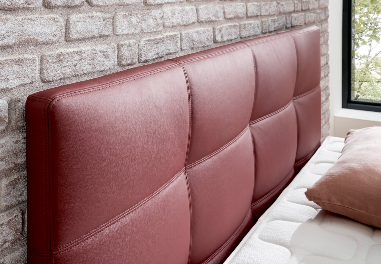 Nahaufnahme eines Boxspringbett-Kopfteil in weinrotem Leder mit gestepptem Design, seitliche Perspektive.