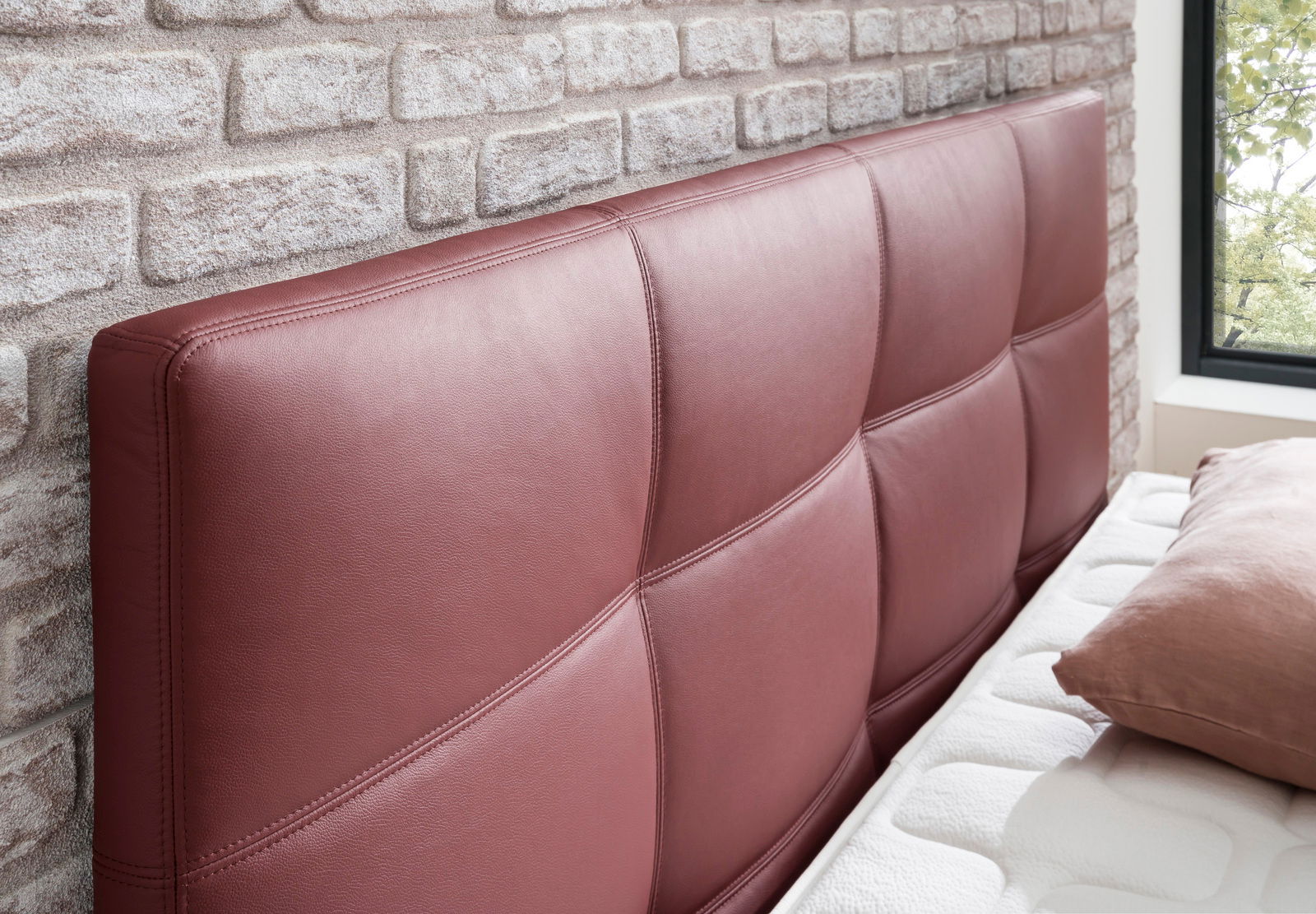 Nahaufnahme eines Boxspringbett-Kopfteil in weinrotem Leder mit gestepptem Design, seitliche Perspektive.