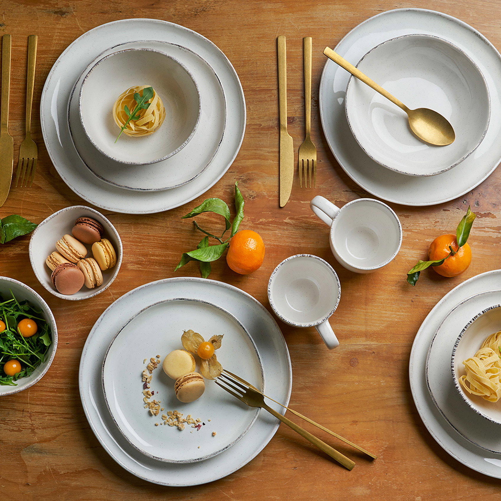 Modernes cremeweißes Geschirr mit goldfarbenem Besteck auf rustikalem Holztisch dekoriert. Stilvolle Tischdekoration mit Pasta, Macarons und frischen Mandarinen