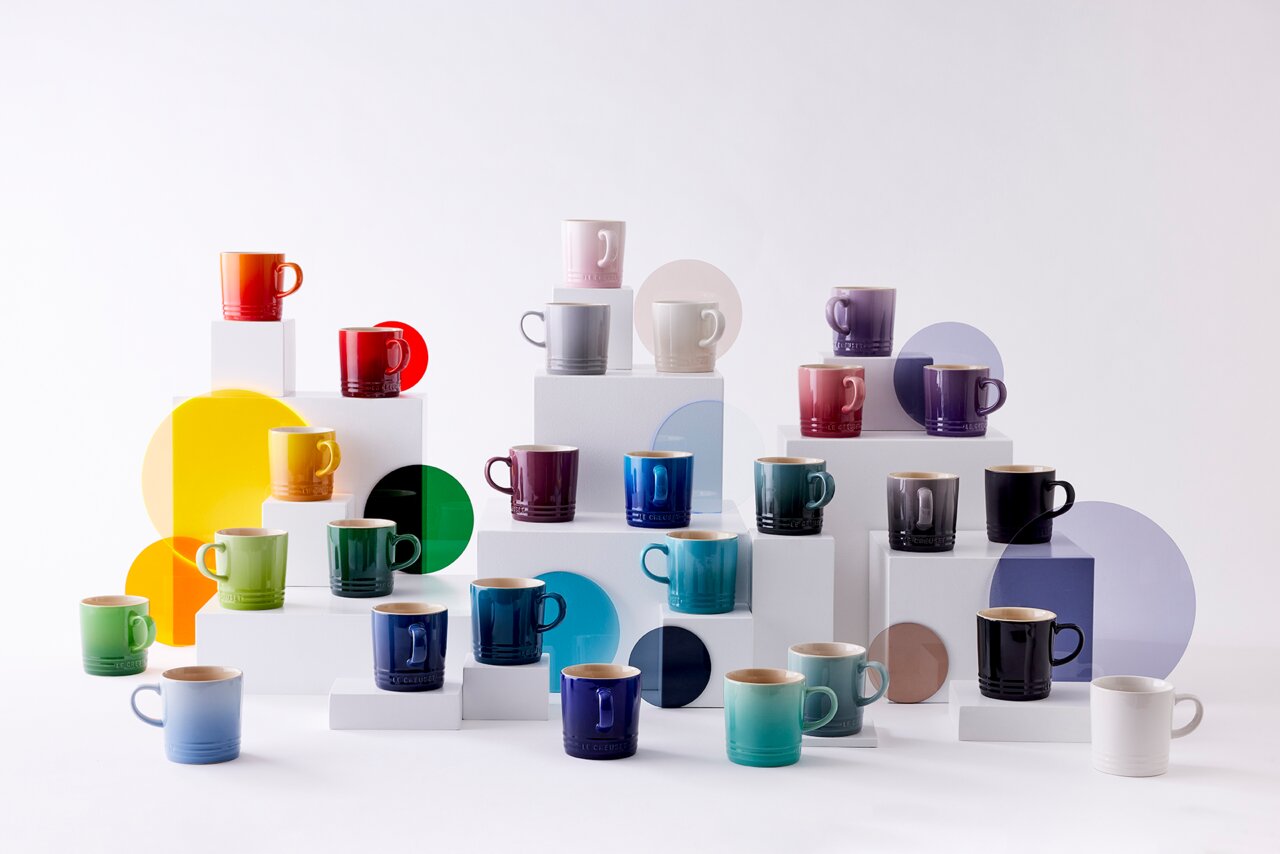 LE CREUSET Becher 0,35l Schwarz Glänzend Verschiedene bunte Becher auf gestuften weißen Podesten, fotografiert aus einer frontalen Perspektive.