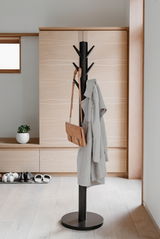 Moderner schwarzer Garderobenständer mit mehreren Haken, auf denen ein Mantel und eine Tasche hängen, in einem hellen Raum mit Holzfußboden und minimalistischer Einrichtung. Seitliche Perspektive.