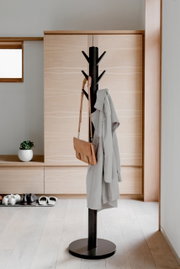 Moderner schwarzer Garderobenständer mit mehreren Haken, auf denen ein Mantel und eine Tasche hängen, in einem hellen Raum mit Holzfußboden und minimalistischer Einrichtung. Seitliche Perspektive.
