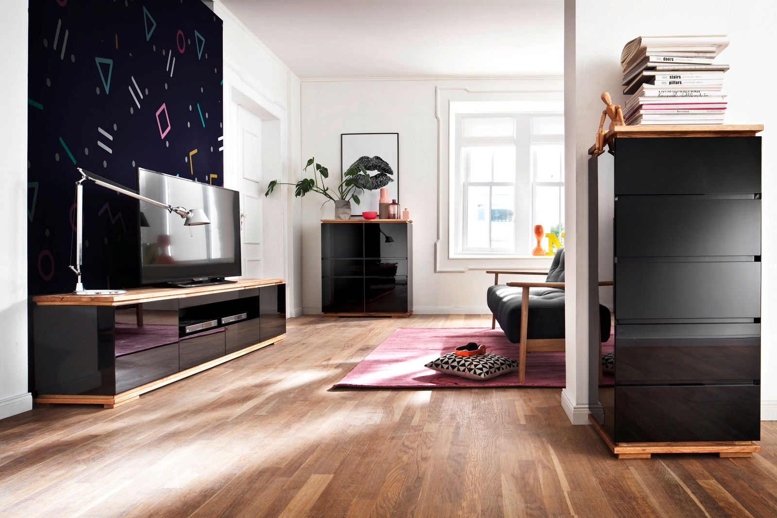 Modernes Wohnzimmer mit TV-Lowboard, Hochkommode und Highboard in schwarzem Hochglanz und Holzoptik, seitliche Perspektive.