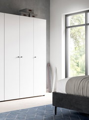 Moderner weißer Kleiderschrank mit drei Türen und schwarzen Knöpfen im hellen Schlafzimmer, frontale Ansicht leicht von links, neben großem Fenster und Bett