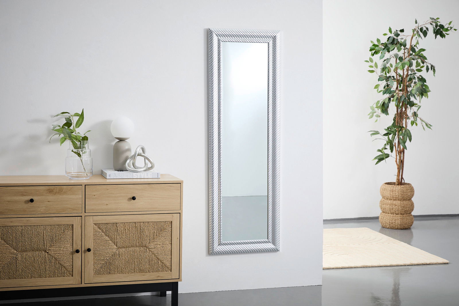 Rechteckiger Wandspiegel mit dekorativem Rahmen, frontal an einer weißen Wand montiert, neben einer Kommode und einer Pflanze.