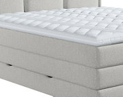 Witte, dik gewatteerde matrastopper met doorgestikt patroon en ritssluiting op een lichtgrijs gestoffeerd boxspringbed, schuin van voren en bovenaf gezien