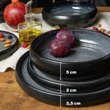 Dreiteiliges graues Tafelservice mit verschiedenen Höhen, präsentiert auf einem Holztisch mit Früchten und Ölflasche, seitliche Perspektive.