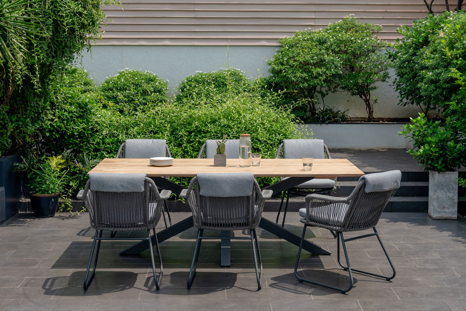 Moderner Gartentisch mit Holzplatte und sechs grauen Stühlen, umgeben von üppigem Grün, aus der Vorderansicht fotografiert.
