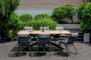 Moderner Gartentisch mit Holzplatte und sechs grauen Stühlen, umgeben von üppigem Grün, aus der Vorderansicht fotografiert.