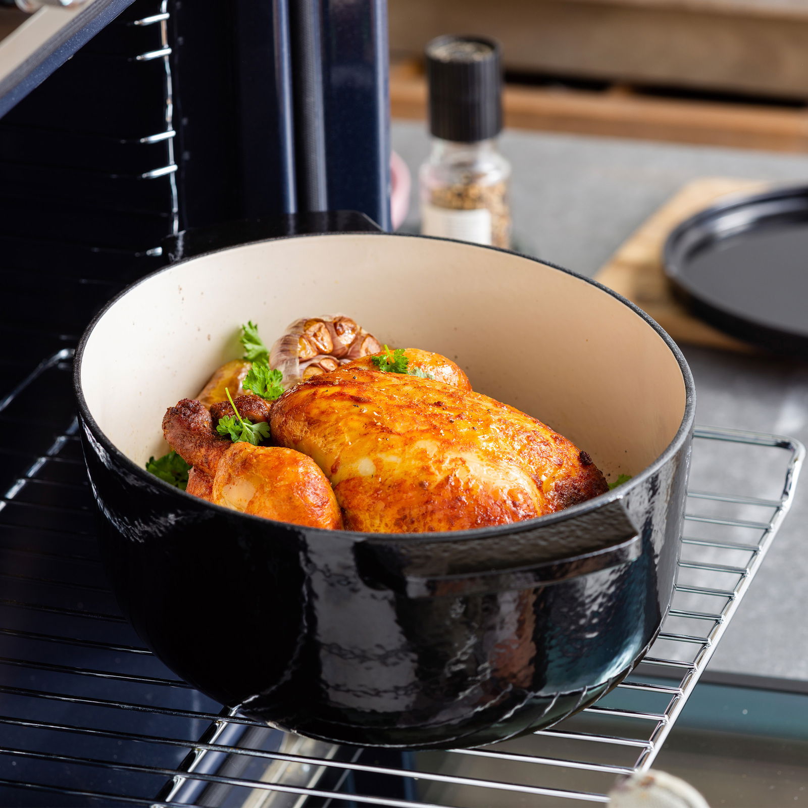 Gusseiserner ovaler Bräter mit gebratenem Hähnchen und Knoblauch, im Ofen auf einem Rost von schräg oben fotografiert.
