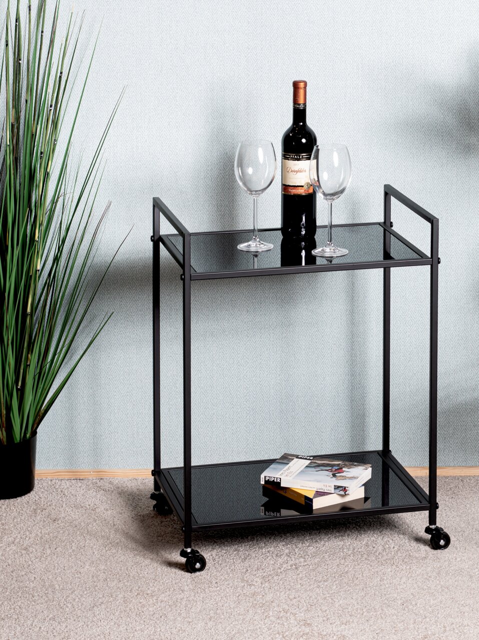 Moderner Beistellwagen mit zwei Ebenen, einer Flasche Wein und zwei Gläsern auf der oberen Ablage, Bücher auf der unteren Ablage, seitliche Perspektive.