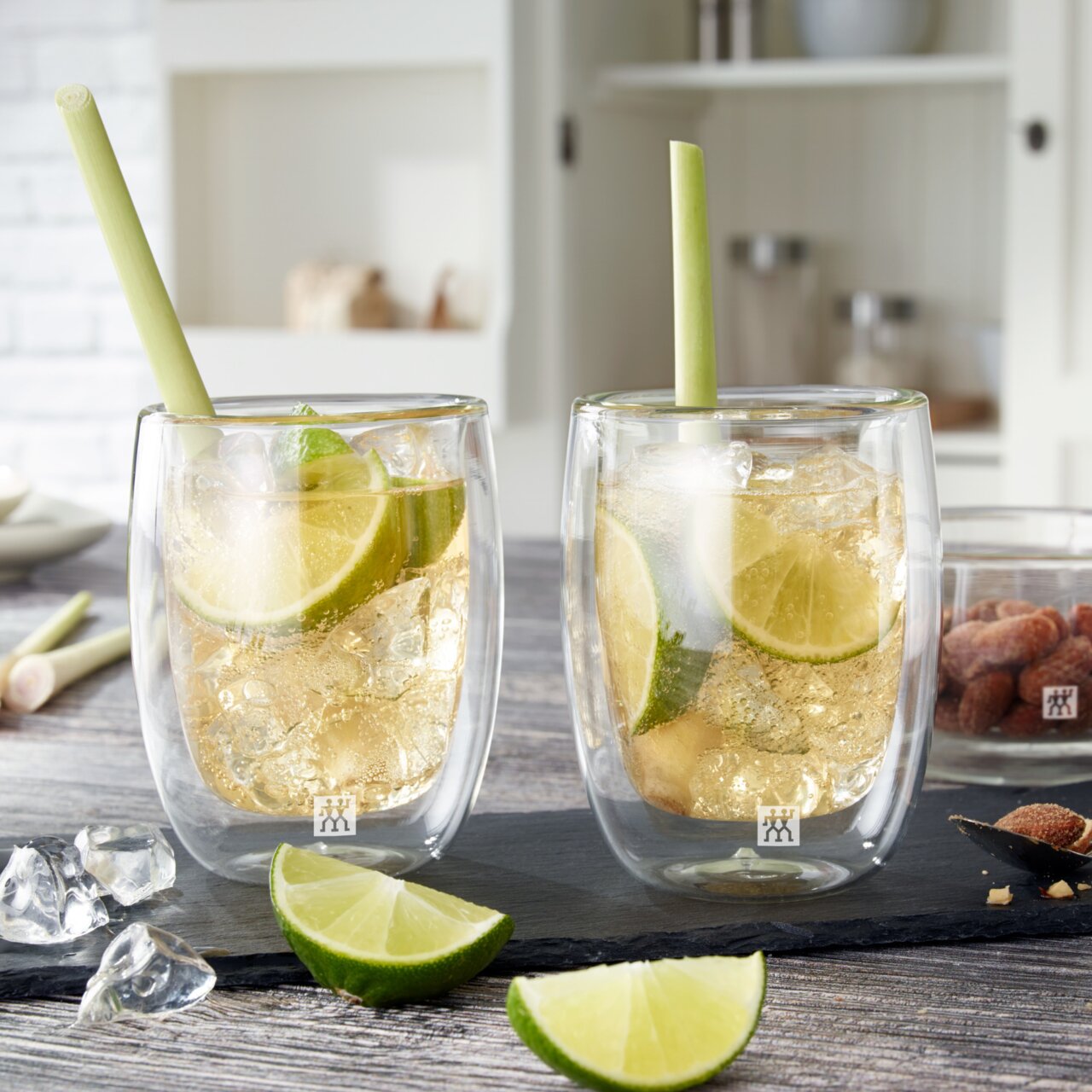 Zwei doppelwandige Kaffeegläser mit Eiswürfeln und Limettenscheiben, gefüllt mit einem erfrischenden Getränk, auf einem Tisch mit Limettenscheiben und Eiswürfeln, aus der Vorderansicht fotografiert.