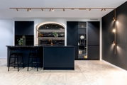 Frontalansicht einer modernen Designküche in Dunkelblau/Schwarz mit geriffelter Front, großer Kücheninsel mit Barhockern, dunkler Stein-Arbeitsplatte, beleuchtetem Rundbogen-Regal und goldfarbener Armatur in einem hellen Raum mit Schienenspots