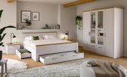 Modernes Schlafzimmer-Set in Weiß mit Holz-Akzenten: Doppelbett mit ausziehbaren Bettkästen, Nachttisch und Kleiderschrank mit Spiegeltüren, gezeigt in schräger Frontansicht im hellen Raum.