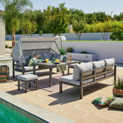 Ess-Lounge-Set auf einer Terrasse mit Blick auf den Pool, bestehend aus einem Tisch, Bänken mit grauen Kissen und einem Sonnenschutz, umgeben von Pflanzen.