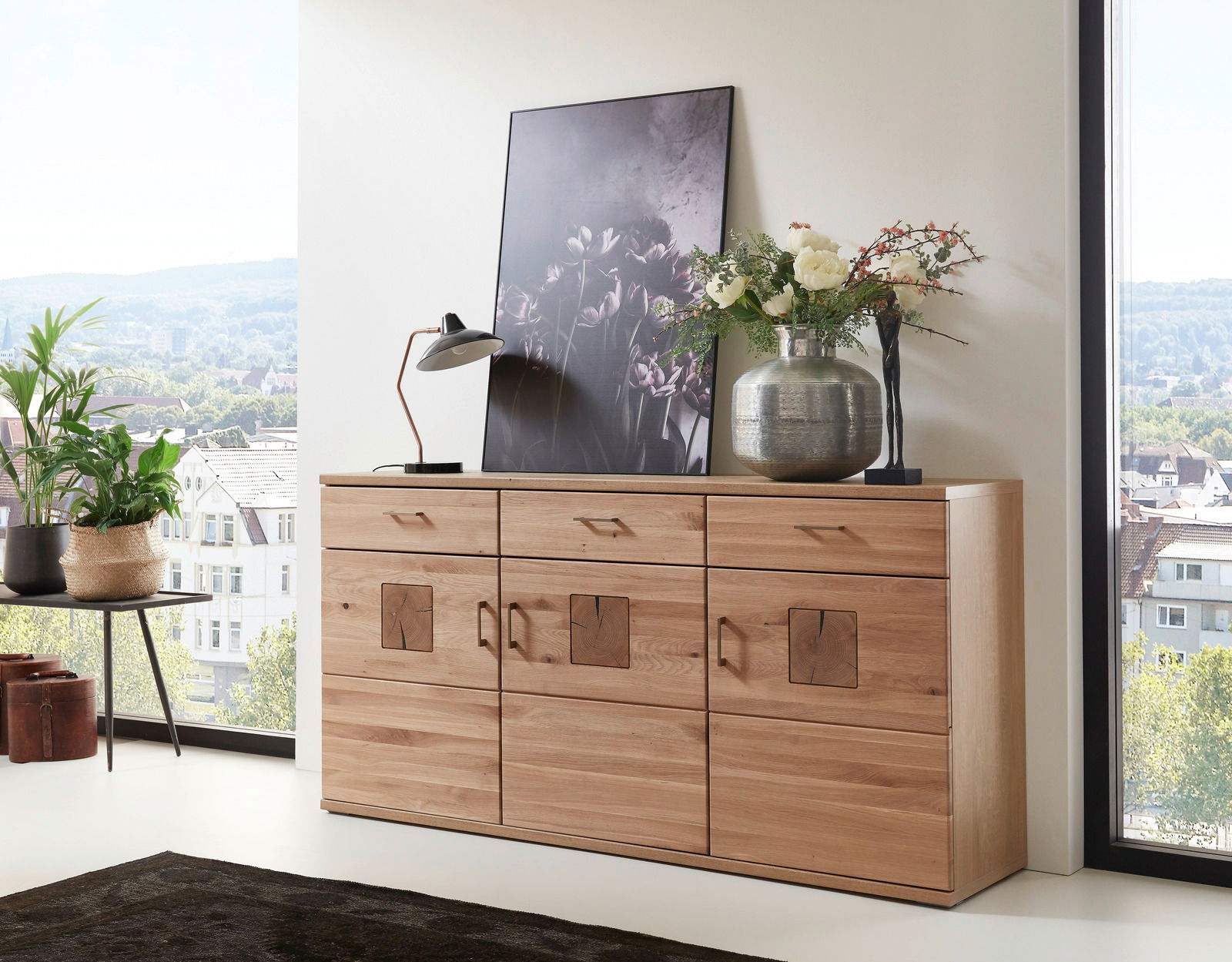 Holz-Sideboard mit drei Schubladen und dekorativen Elementen, darauf eine Lampe, ein Bild und eine Vase mit Blumen, seitliche Perspektive.