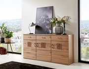 Holz-Sideboard mit drei Schubladen und dekorativen Elementen, darauf eine Lampe, ein Bild und eine Vase mit Blumen, seitliche Perspektive.