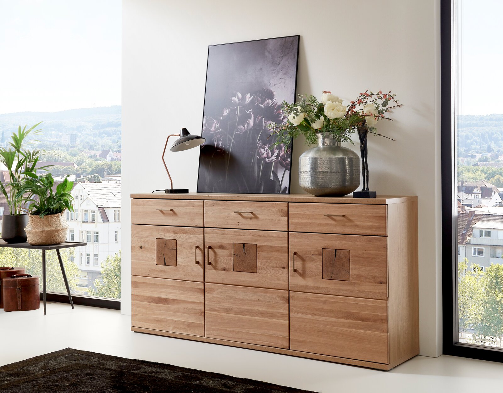 Holz-Sideboard mit drei Schubladen und dekorativen Elementen, darauf eine Lampe, ein Bild und eine Vase mit Blumen, seitliche Perspektive.
