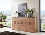 Holz-Sideboard mit drei Schubladen und dekorativen Elementen, darauf eine Lampe, ein Bild und eine Vase mit Blumen, seitliche Perspektive.