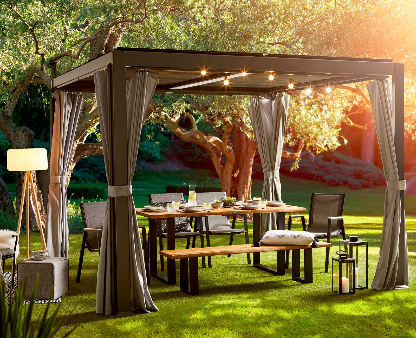 Gartentisch unter einem Pavillon mit Lichterkette, umgeben von Stühlen und Bänken, in einem sonnigen Garten, seitliche Perspektive