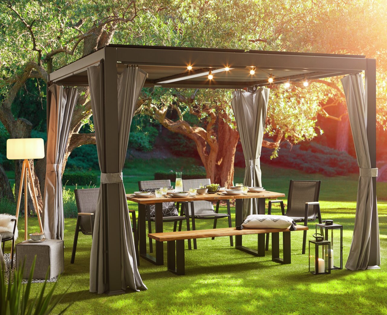 Gartentisch unter einem Pavillon mit Lichterkette, umgeben von Stühlen und Bänken, in einem sonnigen Garten, seitliche Perspektive