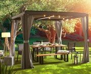 Gartentisch unter einem Pavillon mit Lichterkette, umgeben von Stühlen und Bänken, in einem sonnigen Garten, seitliche Perspektive