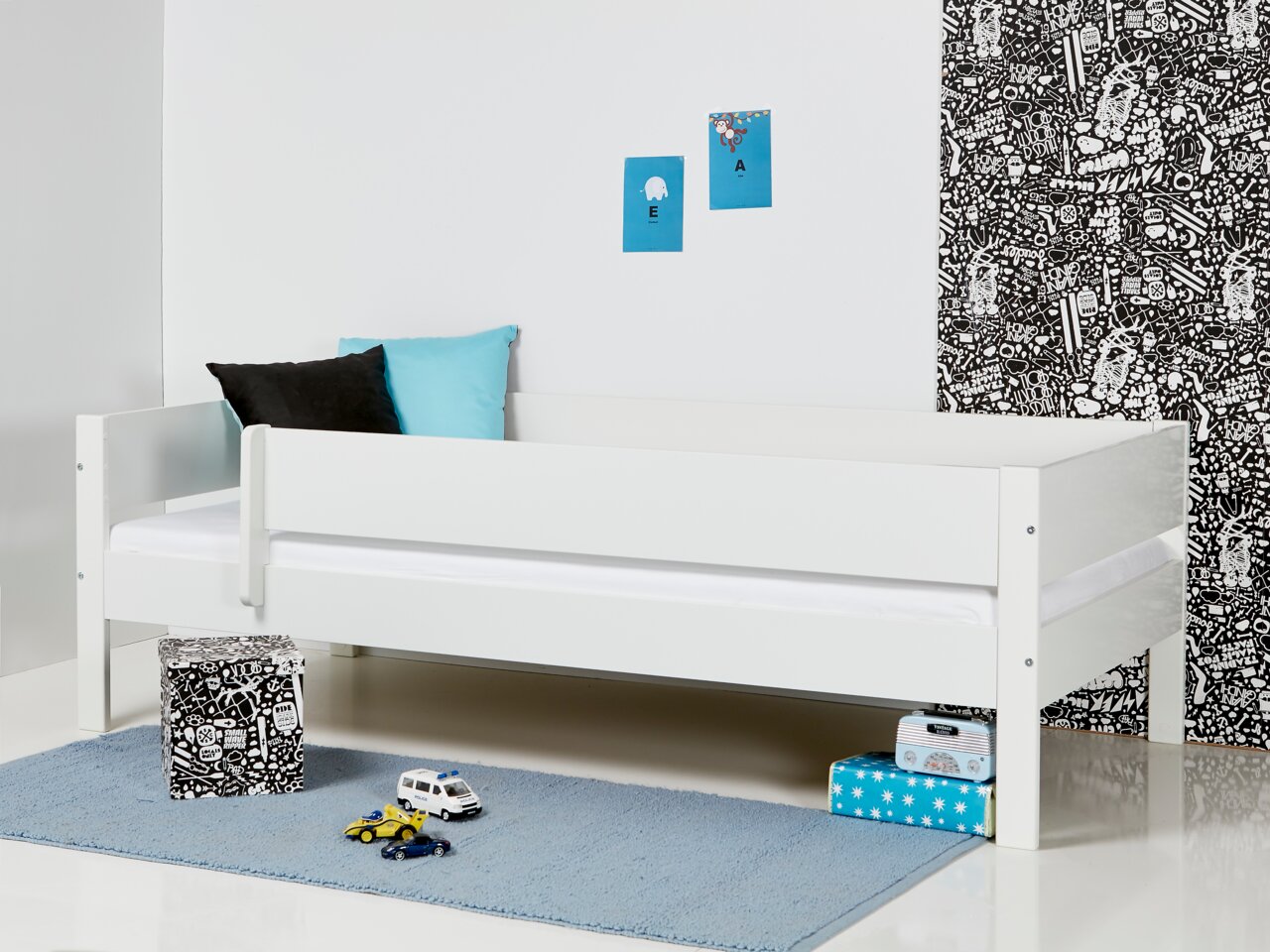 Weißes Kinderbett mit 3/4 Absturzsicherung, seitliche Perspektive, dekoriert mit Kissen und Spielzeug auf einem blauen Teppich.