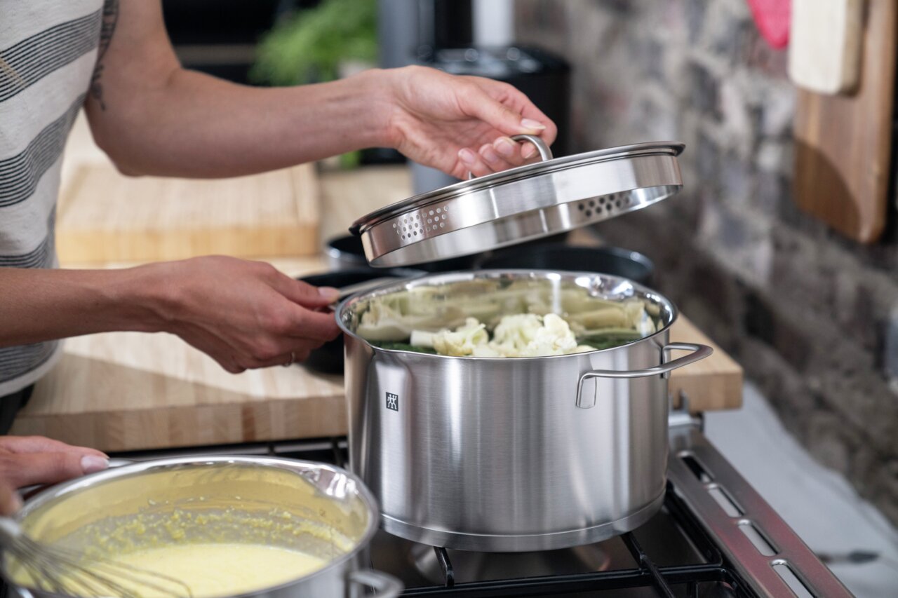 Person hebt den Deckel eines Edelstahltopfs an, der auf einem Herd steht, mit Gemüse im Topf. Seitliche Perspektive.