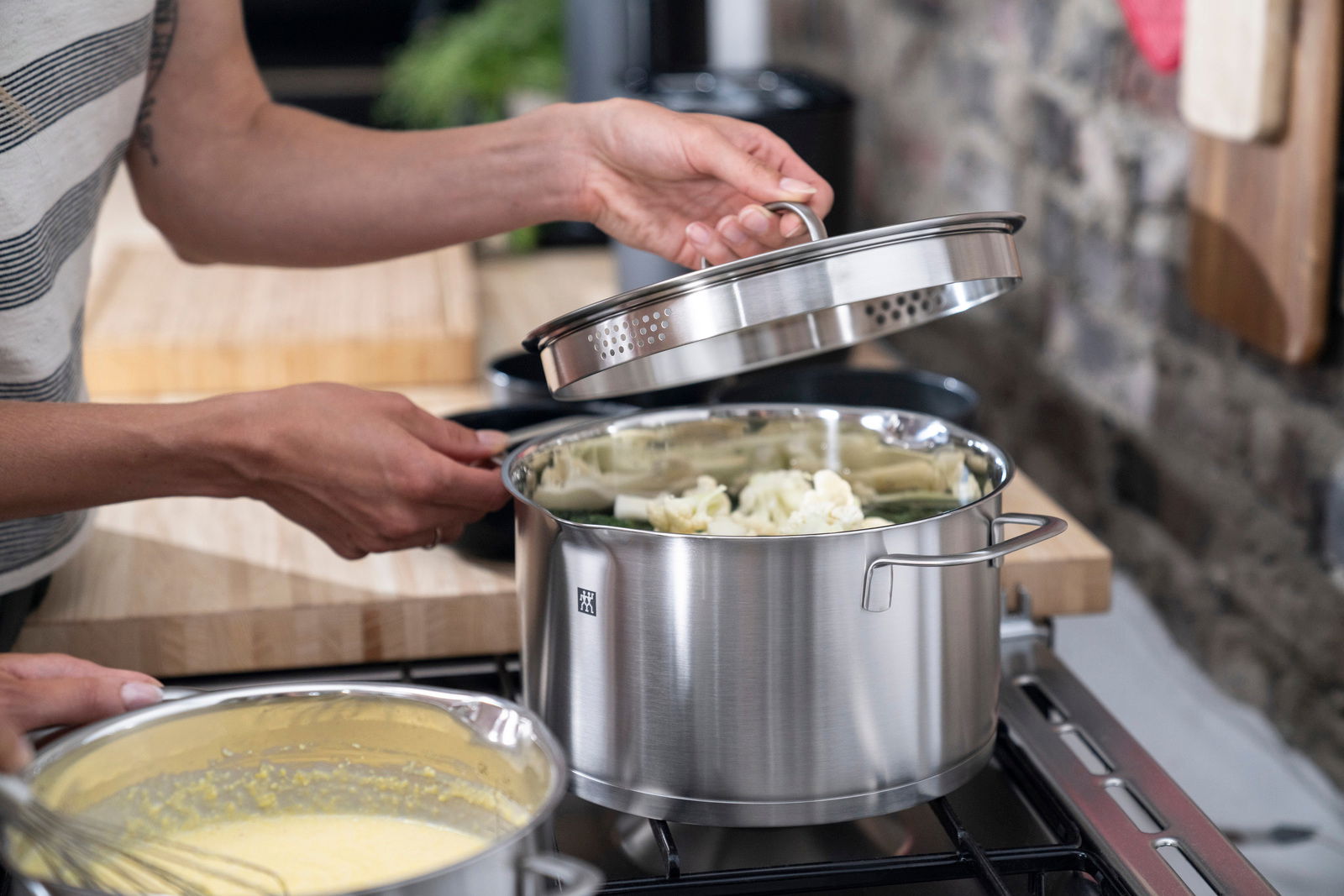 Person hebt den Deckel eines Edelstahltopfs an, der auf einem Herd steht, mit Gemüse im Topf. Seitliche Perspektive.