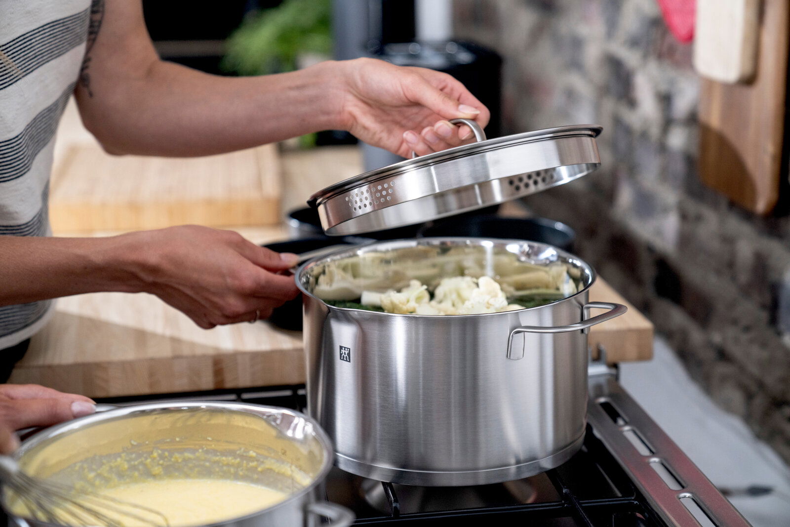 Person hebt den Deckel eines Edelstahltopfs an, der auf einem Herd steht, mit Gemüse im Topf. Seitliche Perspektive.
